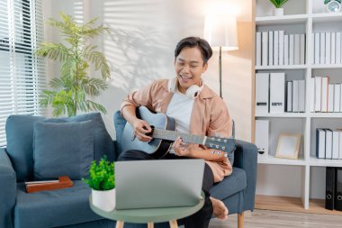 Genç bir adam oturma odasında zevkle gitar çalıyor, akor çalışırken ritmin tadını çıkarıyor. Rahat duruşu ve gülüşü mutluluğu ve müzikal tutkuyu yansıtıyor..