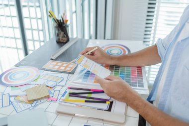 Bir tasarımcı masadaki yaratıcı bir UX UI projesi üzerinde çalışıyor, renk paletleri ve düzen şablonlarını odaklanma ve hassasiyetle düzenliyor, modern tasarım ve yeniliği temsil ediyor.