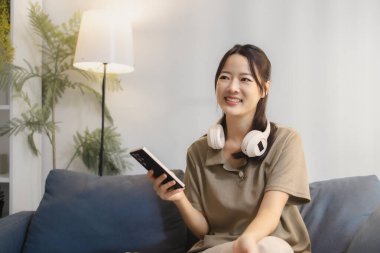 Kanepede oturan genç bir kadın, elinde kulaklıkla elinde akıllı bir telefonla gülümsüyor. Yakınına bir oyun denetleyici yerleştirilir, rahatlamış bir boş zaman anı gösterir.