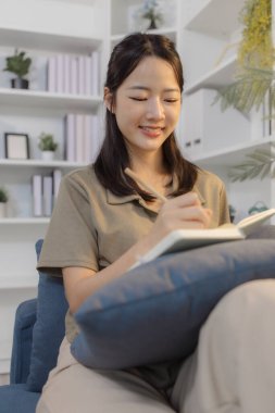 Genç bir kadın, modern bir ofisteki koltukta rahat bir şekilde oturur elinde bir defter tutarak düşünceli bir şekilde yazar. Odaklanmış ve rahat görünüyor, sakin bir kişisel yansımanın tadını çıkarıyor..