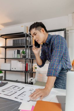 Ofis masasında çalışan kendinden emin genç mimar, ev planları çiziyor ve inşaat detaylarını telefonda tartışıyor, mimari tasarım ve proje planlamasında doğruluk ve yaratıcılığa odaklanıyor