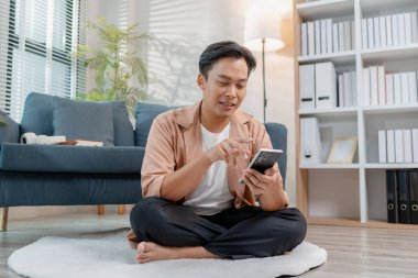 Genç bir adam, rahat bir oturma odasında akıllı telefon kullanırken bacak bacak bacak üstüne atmış, gülümseyerek yerde rahatça oturuyor. Rahatlamış atmosfer konfor, yaşam tarzı ve ev bağlantısını vurguluyor.