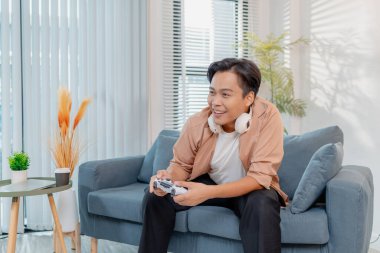 Neşeli bir genç adam evinde bir kanepede oturuyor, elinde bir oyun kumandası tutuyor ve neşe ve heyecanla video oyunları oynuyor, kahkaha ve zaferi ifade ediyor, aynı zamanda oyuna dalmış..