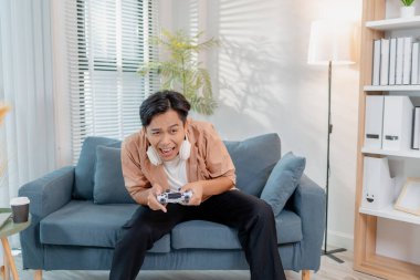 Neşeli bir genç adam evinde bir kanepede oturuyor, elinde bir oyun kumandası tutuyor ve neşe ve heyecanla video oyunları oynuyor, kahkaha ve zaferi ifade ediyor, aynı zamanda oyuna dalmış..