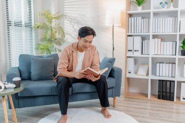 Genç bir adam kanepede rahatça oturup kitap okuyor, düşünceli ve odaklanmış görünüyor. İçerideki rahat ortam rahatlama, konsantrasyon ve bilgiyi ön plana çıkarıyor..