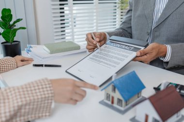 Bir emlakçı ve müşteri emlak satın alma seçeneklerini tartışıyor, sözleşmeyi gözden geçiriyor, kredi şartlarını hesaplıyor ve ev modellerini konut yatırımı kararının bir parçası olarak değerlendiriyor..