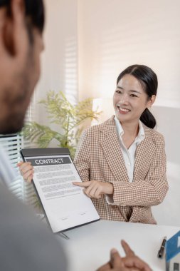 Bir emlakçı, şartları açıklamak için ev modelleri ve hesap makinesi kullanarak bir müşteriyle emlak sözleşmelerini tartışır. Sahne, müzakere, karar verme ve mali planlamayı vurguluyor.