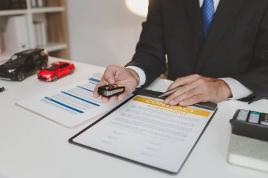 Resmi takım elbiseli bir işadamı, araba sigortası detaylarını, küçük kırmızı bir araba modelini elinde tutarak ve sözleşme belgelerine işaret ederek, anlaşmayı, korumayı ve mali planlamayı sembolize ederek açıklıyor.