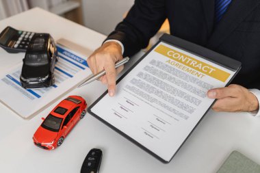 Takım elbiseli bir işadamı, masasında minyatür araba modelleri olan araç sigortası, finansal planlama ve yasal anlaşmaları sembolize eden araba sigortası ve sözleşme belgeleri sunuyor..