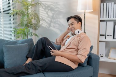 Genç bir adam akıllı telefonunu kullanırken gülümseyerek evde dinlenir. Boynunda kulaklıklarla boş zamanlardan zevk alıyor, rahat bir şekilde oturuyor ve modern bir oturma odasında rahat hissediyor..