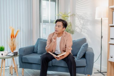 Genç bir adam kanepede rahatça oturup kitap okuyor, düşünceli ve odaklanmış görünüyor. İçerideki rahat ortam rahatlama, konsantrasyon ve bilgiyi ön plana çıkarıyor..