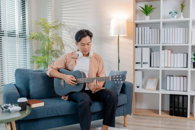 Genç bir adam oturma odasında zevkle gitar çalıyor, akor çalışırken ritmin tadını çıkarıyor. Rahat duruşu ve gülüşü mutluluğu ve müzikal tutkuyu yansıtıyor..