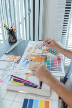 Odaklanmış bir tasarımcı UX / UI tasarım fikirleri üzerinde çalışıyor, bir tahtaya tutturulmuş renk paletleri ve arayüz yapılarını analiz ediyor. Sahne yaratıcılık, planlama ve dijital yeniliği yansıtıyor.