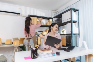 İki genç kadın, kargo kutuları ve belgelerle çevrili küçük bir iş ofisinde birlikte çalışıyorlar. Siparişleri tartışıyorlar, paketleri hazırlıyorlar ve envanteri bilgisayarda yönetiyorlar..