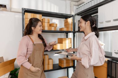 İki genç kadın, küçük bir işletme ofisinde birlikte çalışıyorlar, etrafları paket ve belgelerle çevrili. Görevleri tartışıyor, envanterleri kontrol ediyor ve takım çalışması ve odaklanmayla paketleri hazırlıyorlar..