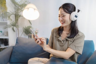 Genç bir kadın rahat bir kanepede kulaklıklarla oturuyor, elinde akıllı bir telefonla gülümsüyor ve müzik dinliyor. Evde teknoloji ve eğlenceyi birleştirerek rahatlatıcı bir anın tadını çıkarıyor..