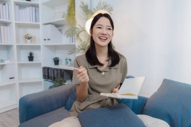 Genç bir kadın, modern bir ofisteki koltukta rahat bir şekilde oturur elinde bir defter tutarak düşünceli bir şekilde yazar. Odaklanmış ve rahat görünüyor, sakin bir kişisel yansımanın tadını çıkarıyor..