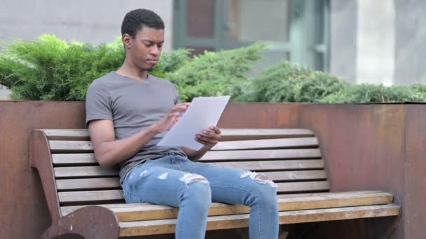 Giovane uomo africano lettura documenti seduta su panchina — Video Stock