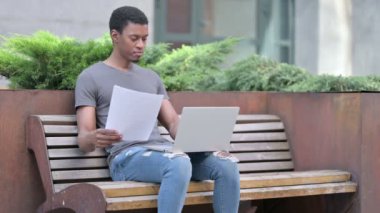 Genç Afrikalı Adam Bench 'te kağıt ve dizüstü bilgisayar üzerinde çalışıyor. 
