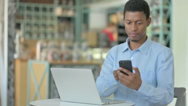 Ung afrikansk man som använder smartphone och laptop i Cafe — Stockvideo