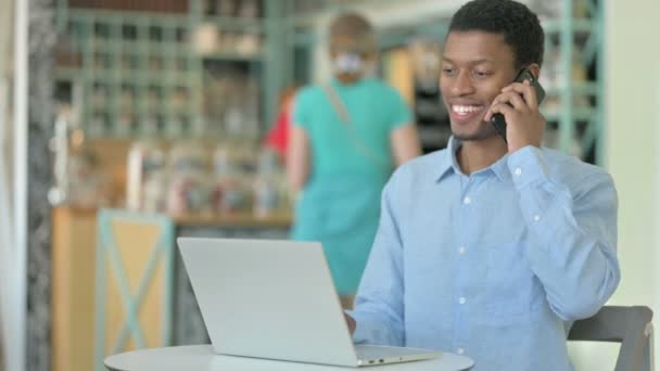 Ung afrikansk man med bärbar dator Talar på smartphone i Café — Stockvideo