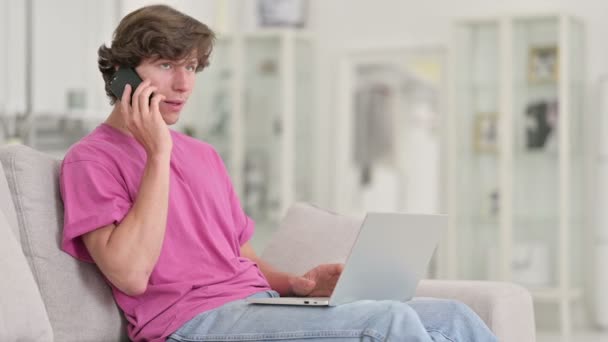 Joven hombre casual con el ordenador portátil hablando en el teléfono inteligente en casa — Vídeos de Stock