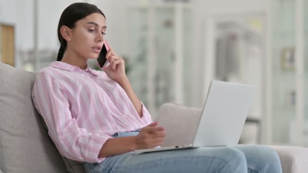 Serieuze Latijnse vrouw met laptop praten op smartphone thuis — Stockvideo