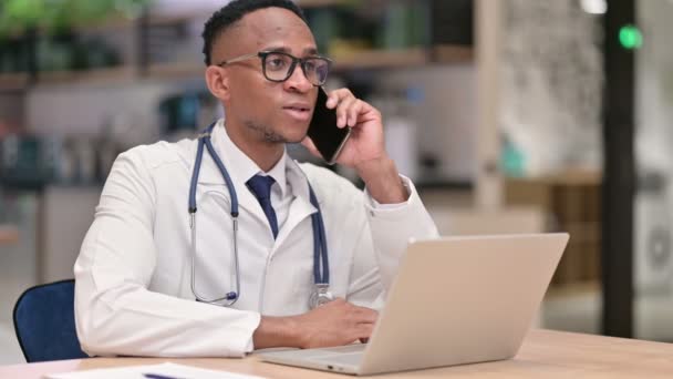 Afrikaanse mannelijke arts met laptop praten op Smartphone in Office — Stockvideo