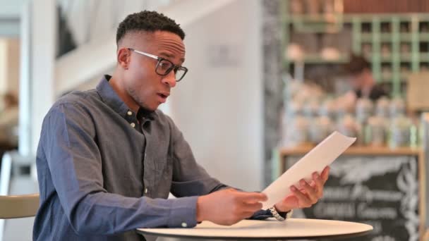 Удивленный молодой африканский человек читает документы в кафе — стоковое видео