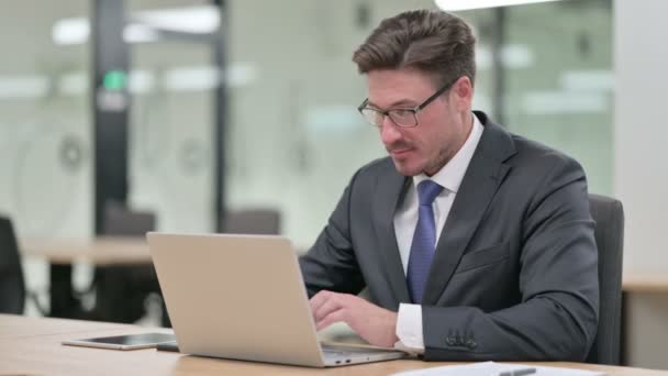 Бизнесмен среднего возраста с ноутбуком улыбается в камеру в офисе — стоковое видео