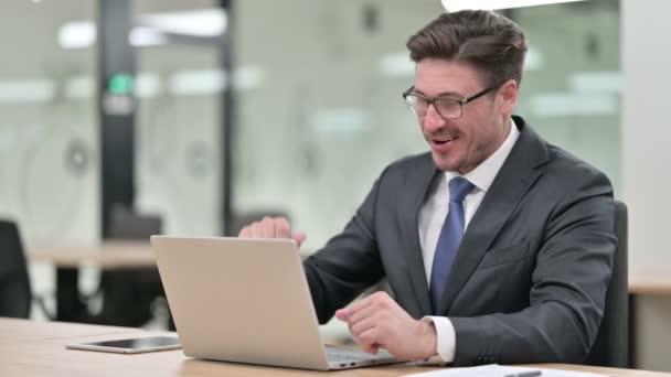 Весёлый бизнесмен среднего возраста разговаривает в видеочате на ноутбуке в офисе — стоковое видео