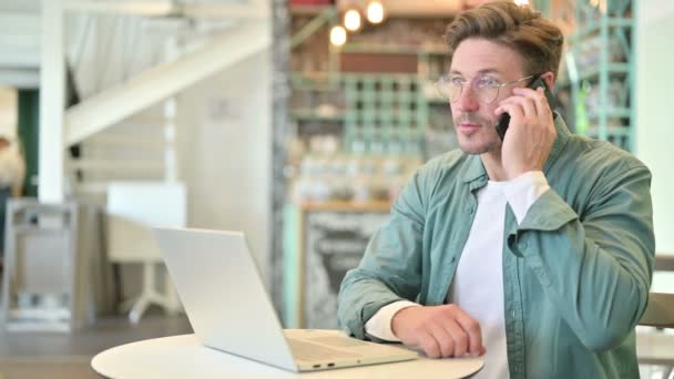 Hombre de mediana edad con el ordenador portátil hablando en el teléfono inteligente en Café — Vídeos de Stock