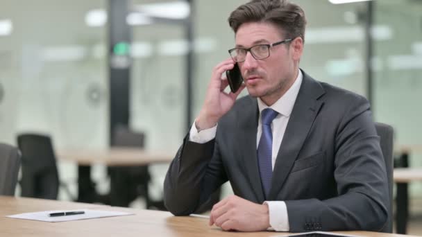 Alegre hombre de negocios de mediana edad que habla en el teléfono inteligente en la oficina — Vídeos de Stock