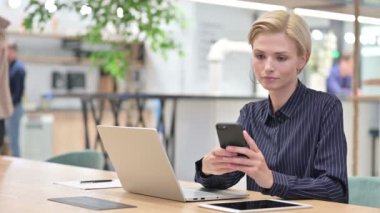 Dizüstü bilgisayarlı genç iş kadını Ofiste Smartphone ile konuşuyor 
