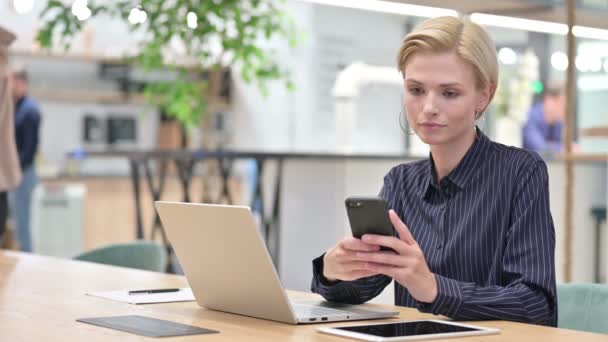 Молодая предпринимательница с ноутбуком разговаривает на смартфоне в офисе — стоковое видео