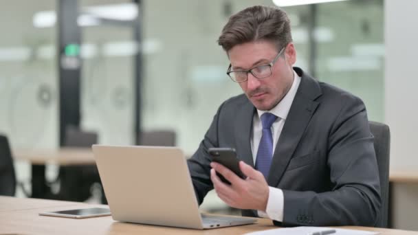 Middle Aged Businessman med Laptop ved bruk av Smartphone i Office – stockvideo