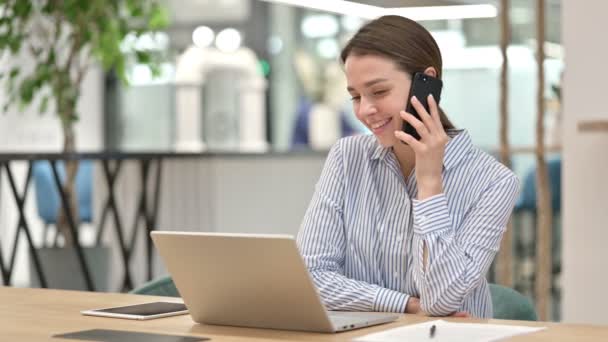 Молодая женщина с ноутбуком разговаривает на смартфоне в офисе — стоковое видео
