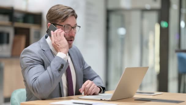 Бизнесмен с ноутбуком разговаривает на смартфоне в офисе — стоковое видео