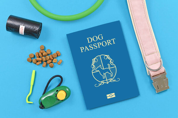 Concept for travelling with dogs showing made up blue dog passport next to pet supplies like collar, tick tweezers, clicker, dog treats or poop bags on blue background