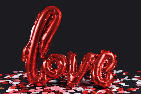 Shiny red balloon forming the word \'love\' on dark black background with colorful confetti on ground