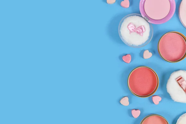 Various pink blush beauty products and powder puffs with ribbons and heart shaped pressed powder on side of blue background with copy space