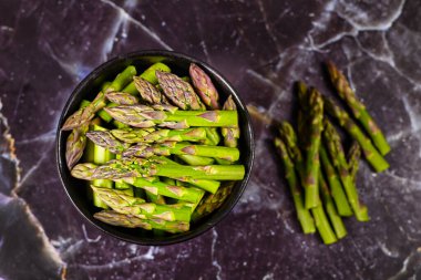Koyu arkaplanda pişirme hazırlıkları sırasında kesilmiş çiğ yeşil kuşkonmaz parçalarıyla kasenin en üst görüntüsü