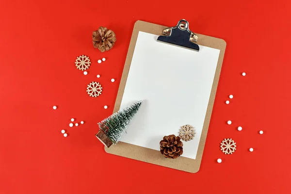 Christmas flat lay with empty clipboard surrounded by small Christmas trees, pine cones, snowballs and snowflake ornaments on red background