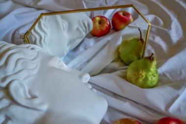 Antik Yunan beyaz heykel alçı mermer mermer kafası altın çerçeveli bir aynaya yansıyan yeşil armut ve kırmızı elma katlı beyaz kumaş bir arka plan