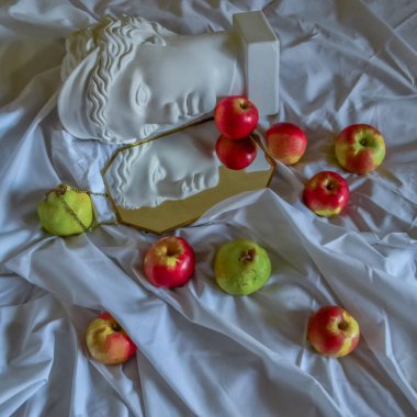 Antik Yunan beyaz heykel alçı mermer mermer kafası altın çerçeveli bir aynaya yansıyan yeşil armut ve kırmızı elma katlı beyaz kumaş bir arka plan