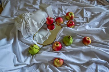 Antik Yunan dönemine ait beyaz heykel alçı mermer mermer kafası, altın çerçeveli bir aynaya, yeşil armutlar ve kırmızı turuncu elmalarla kaplı beyaz kumaştan oluşan bir arka plana yansıyor.