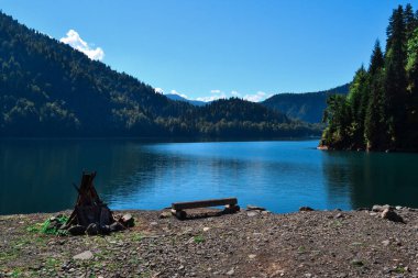 Güzel, mavi, berrak zümrüt, güneşli göl, yeşil ağaçların arasında parlıyor. Yüksek dağlar, şenlik ateşi ve sahil şeridinde oturma tezgahı.