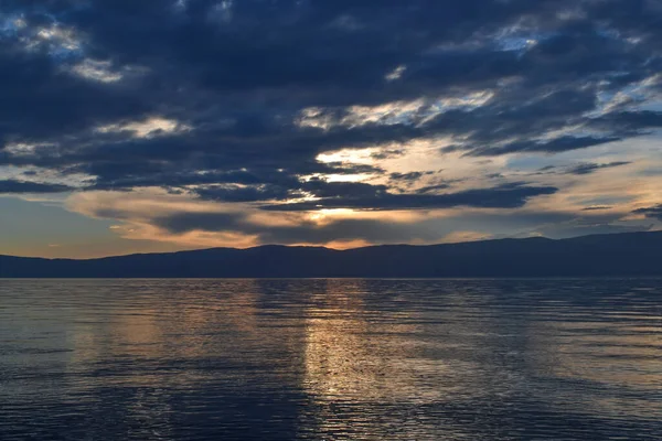 stock image view of the clear calm undulating blue water of Lake Baikal, mountains on the horizon, sunset sky, clouds, reflection