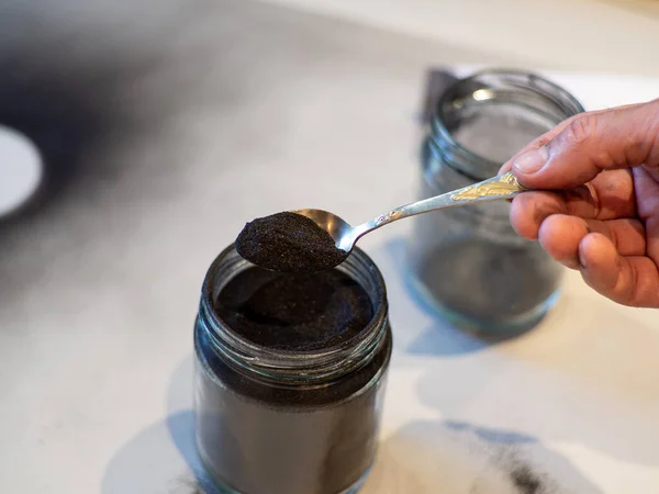 Homemade face mask and scrub with activated powder in glass jar on white background, iron spoon with charcoal