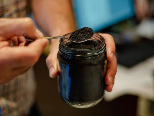 Homemade face mask and scrub with activated powder in glass jar on white back, iron spoon with coal